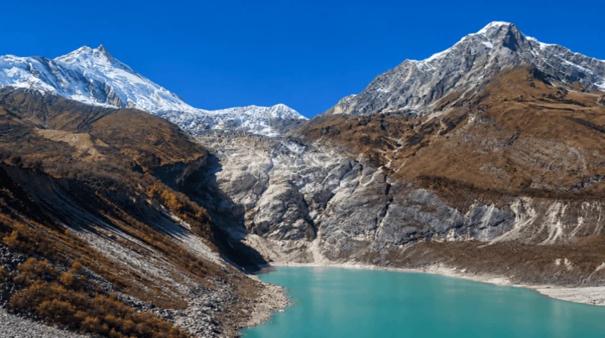 Birendra Taal on the Manaslu Circuit Trek route, featuring a turquoise glacial lake beneath towering snow-capped Himalayan peaks and rugged rocky slopes under a clear deep-blue sky in Nepal's Manaslu region.