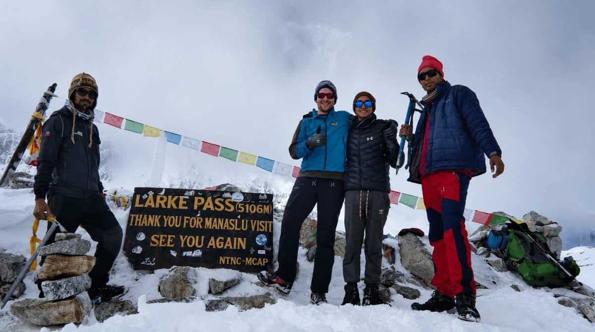 Trekkers with Thrill Himalaya Treks standing at the snowy Larkya La Pass summit (5,106m) decorated with colorful Buddhist prayer flags on the Manaslu Circuit Trek in Nepal.